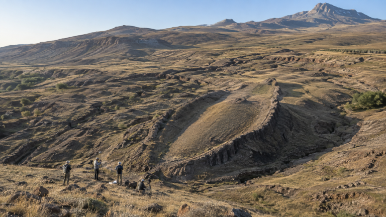 Ilustração de pesquisadores observando formação rochosa alongada em paisagem montanhosa da Turquia