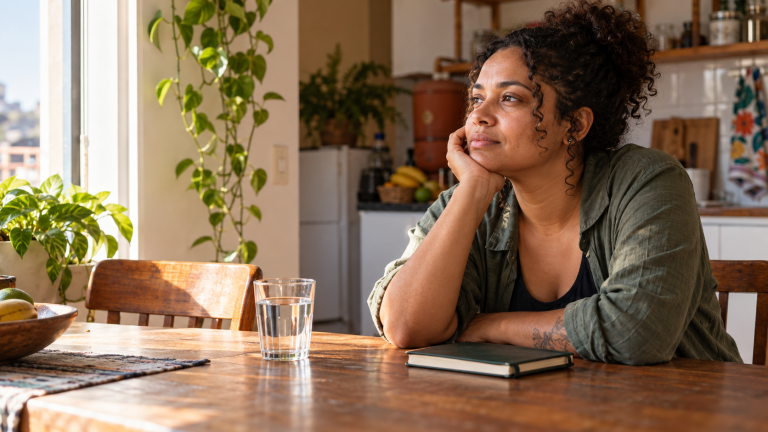 Dia Mundial da Saúde Mental: até descansar virou motivo de culpa? Mulher sentada à mesa em uma pausa silenciosa, em casa, com luz do dia